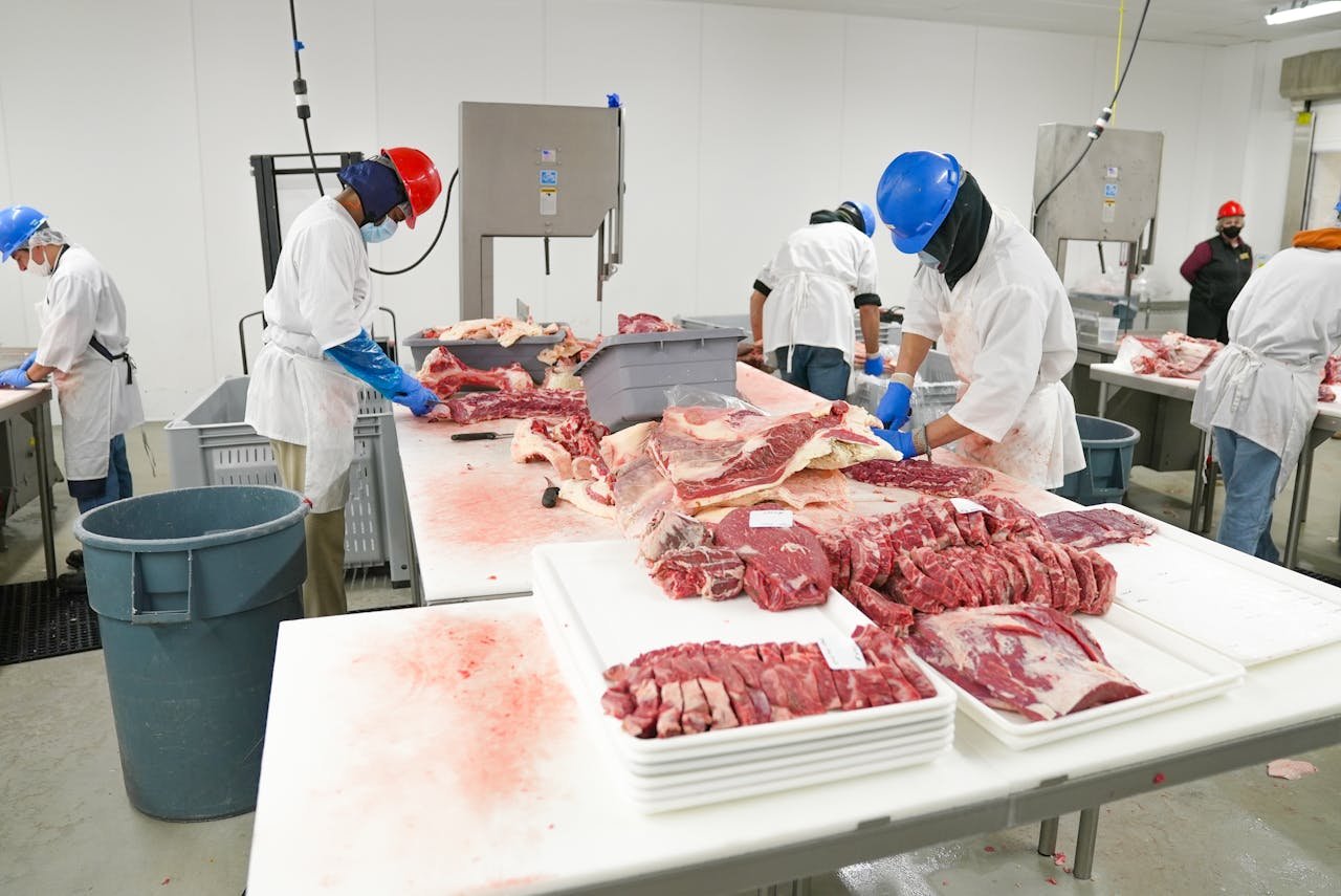 why-choose-us-02 Workers in a meat processing facility wearing safety gear and cutting meat indoors.